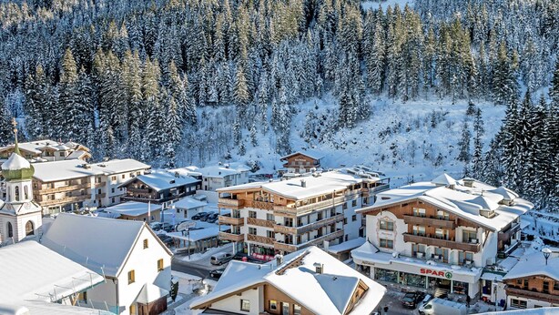 Un pueblo en la montaña cubierto de nieve con edificios de varios pisos, incluyendo un hotel con balcones y una estación de esquí al fondo.
