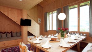 Un intérieur en bois comprenant une table à manger pour quatre personnes, un canapé avec des coussins, un téléviseur fixé au mur et de grandes fenêtres avec des rideaux.