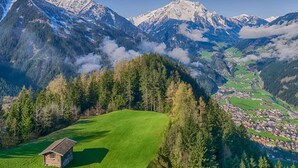 Un paysage de montagne avec une petite cabane en bois sur un pré verdoyant, un village en contrebas et des sommets enneigés en arrière-plan.