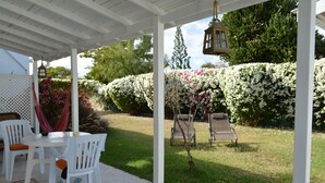 An outdoor patio with white chairs and a table, surrounded by lush greenery and flowering bushes. -  Tropical Gecko Studio - Only 80 meters from the Ocean! (Silver Sands)