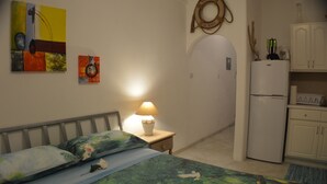 A bedroom with a bed, a nightstand with a lamp, a refrigerator, and a kitchen area with cabinets. -  Tropical Gecko Studio - Only 80 meters from the Ocean! (Silver Sands)