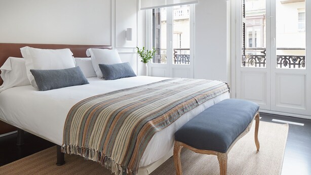 A neatly made bed with a striped blanket and pillows, a wooden bench, and a view of a balcony with a railing. - Pitipombo by FeelFree Rentals (Donostia)