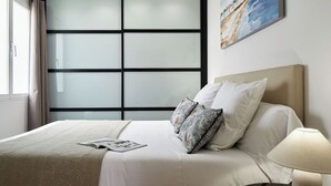 Ein Hotelzimmer mit großem Fenster, einem Bett mit weißen Leinentüchern, gemusterten Kissen, einer Zeitschrift auf dem Bett und einem Gemälde an der Wand.
