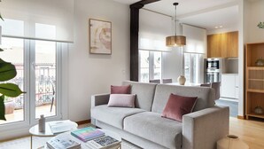 Ein modernes Wohnzimmer mit einem grauen Sofa, einem runden Couchtisch mit Büchern, einem großen Fenster und einer Küche im Hintergrund.