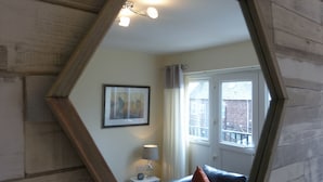 A room with a large mirror reflecting a window, a framed picture on the wall, a small table with a lamp, and a dark-colored sofa with cushions. - The Sawmills (Carlisle)