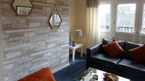 A living room with a modern design, featuring a wooden wall, a black leather sofa with orange cushions, a glass coffee table, and a small side table with a lamp. - The Sawmills (Carlisle)