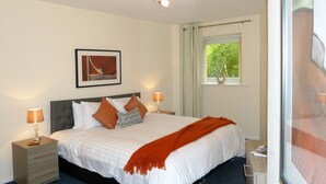 A neatly made bed with a white comforter and orange throw, a bedside lamp, a nightstand with drawers, a framed picture on the wall, and a window with a view of greenery. - The Sawmills (Carlisle)