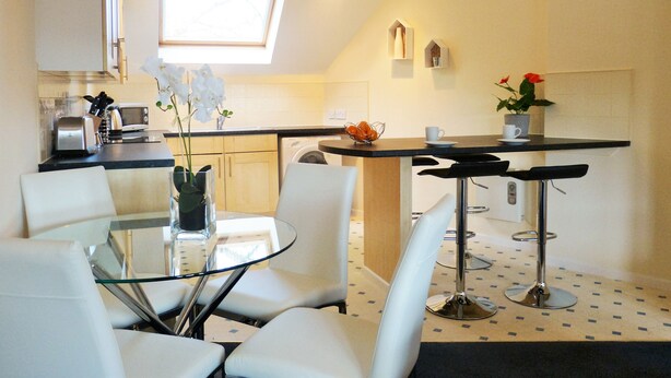 A compact kitchen with a dining area, featuring a glass table, white chairs, and a small bar area. - The Sawmills (Carlisle)