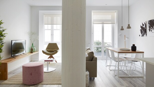 A modern living room with a wooden floor, a pink ottoman, a beige armchair, a dining table with white chairs, and a large window. - Concha Bay 3 by FeelFree Rentals (Donostia)