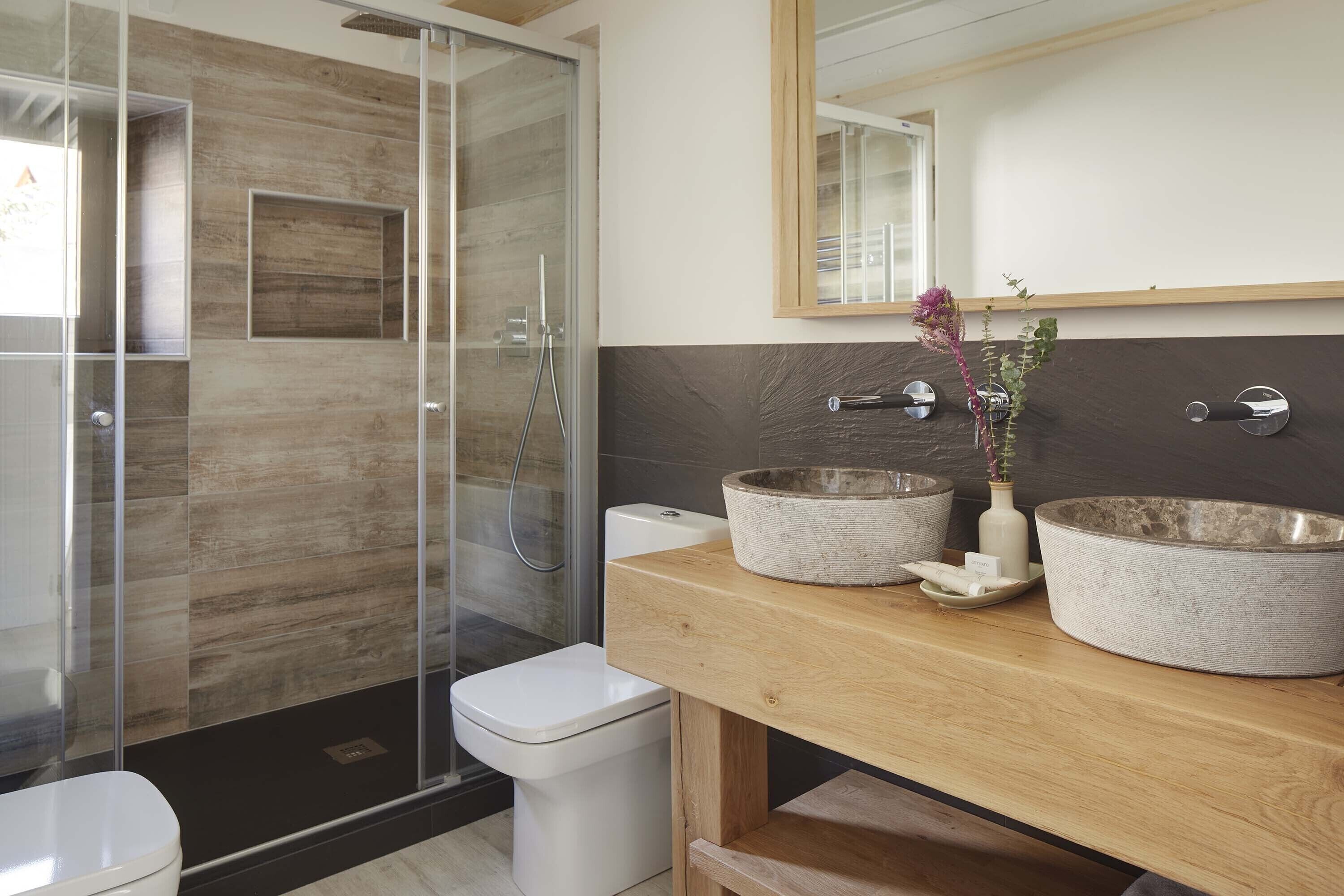 A modern bathroom with a glass shower enclosure, a white toilet, and two stone sinks with a wooden vanity.