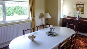 Un comedor con una mesa blanca, sillas, un radiador, una ventana con cortinas, una lámpara y un aparador con un marco de fotos.