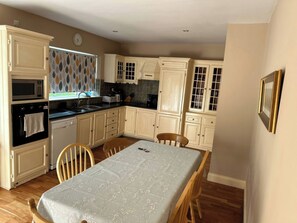 A kitchen with wooden cabinets, a dining table, and a window with leaf-patterned curtains. - The Sanctuary (Churchfield)