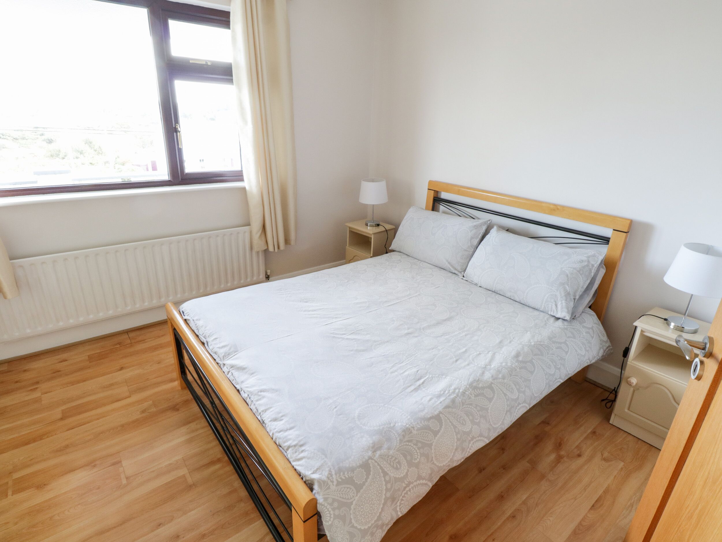 A neatly arranged bedroom with a wooden bed, a bedside table with a lamp, and a window with curtains.