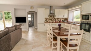 Una cocina moderna con armarios blancos, un comedor con mesa y sillas de madera, horno empotrado y televisor en la sala.