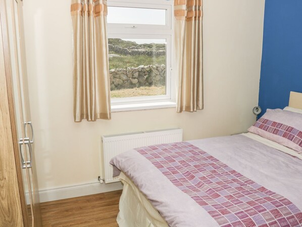 Una camera da letto con un letto, una finestra con tende, un termosifone e vista su un muro in pietra e vegetazione all'esterno.