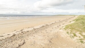 A wide sandy beach with gentle waves, a grassy dune, and a cloudy sky.