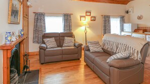 A cozy living room with a fireplace, two sofas, wooden flooring, and a kitchen area in the background. - Rose Cottage (Ramsgrange)