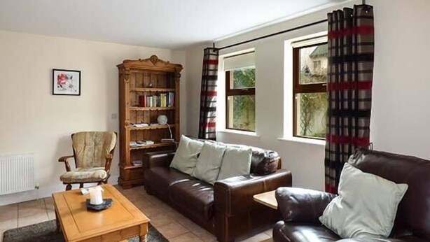 Un salon avec un canapé en cuir marron, une table basse en bois, une bibliothèque en bois et une fenêtre ornée d’un rideau à carreaux.