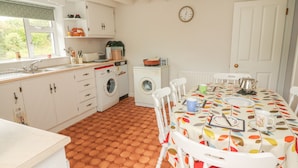A kitchen with white cabinets, a washing machine, a dining table with a colorful leaf-patterned tablecloth, and a clock on the wall. - Limestone Cottage (Carran)