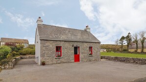 A stone cottage with a red door and two windows, surrounded by a gravel driveway and a stone wall. - Limestone Cottage (Carran)