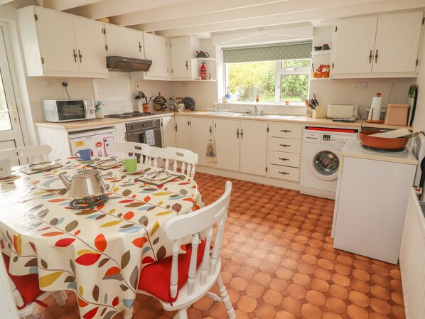 Une cuisine avec des meubles blancs, une table à manger recouverte d’une nappe colorée, un lave-linge et une fenêtre donnant sur des arbres.