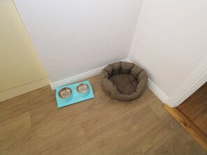 Un rincón de una habitación con piso de madera, una cama para mascotas color café y dos cuencos metálicos sobre un tapete azul.