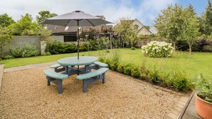 A patio area with a round table and benches, surrounded by gravel, with a house and garden in the background. - 2 Castle Cove (Dromahair)