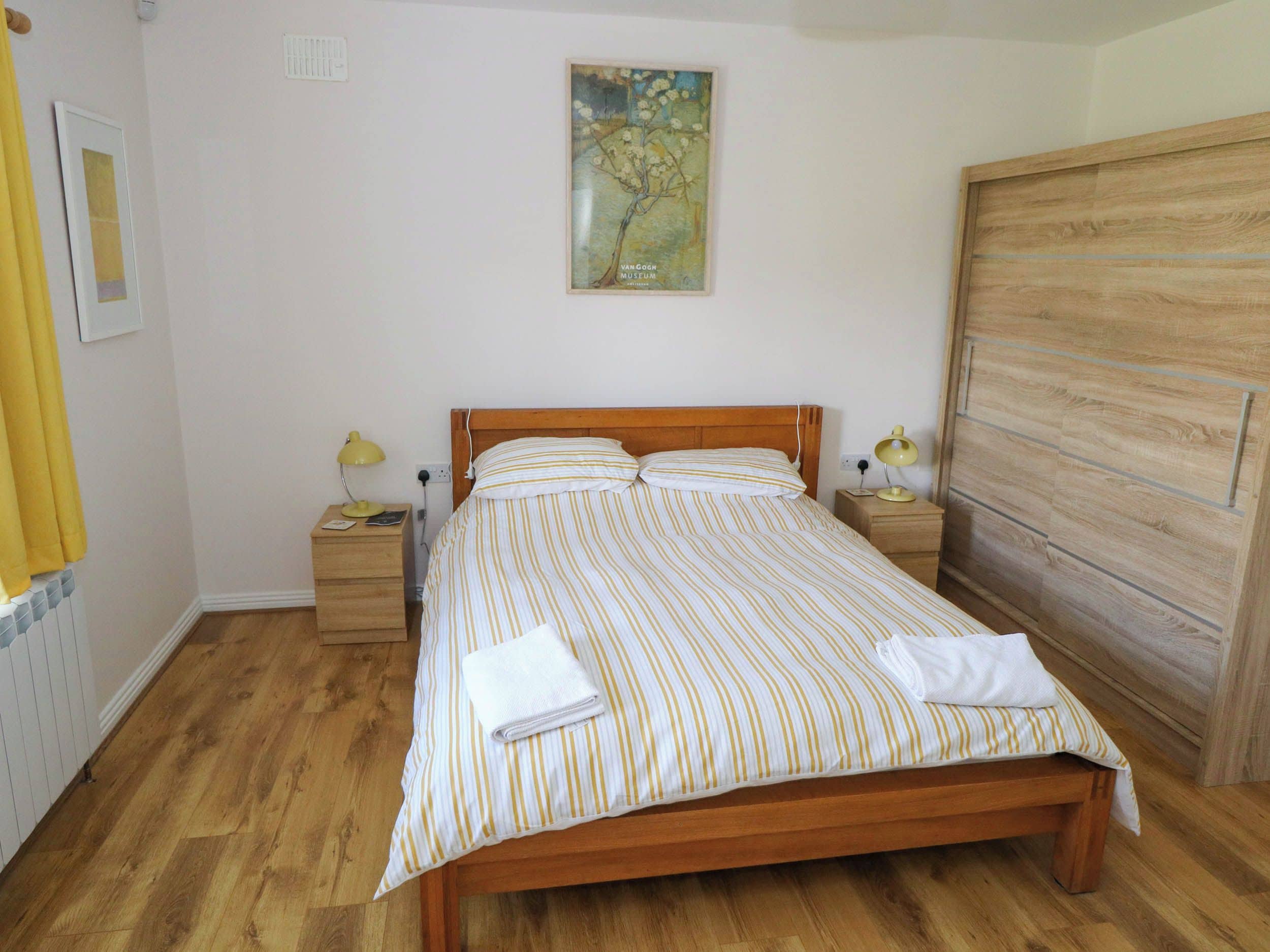 Una cama bien hecha con ropa de cama a rayas, dos lámparas de noche, un cabecero de madera, un armario de madera y un cuadro en la pared.