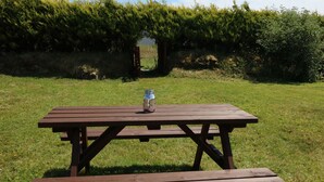 Una mesa de picnic de madera con un tarro encima, ubicada en un campo cubierto de césped y un seto al fondo.