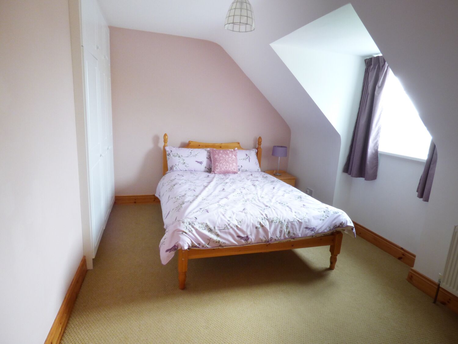Quarto com teto inclinado, cama de madeira, mesa de cabeceira, abajur e janela com cortinas.