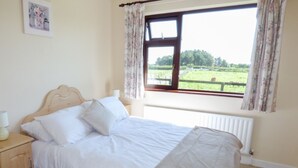 Een slaapkamer met een houten bed, wit beddengoed, een nachtkastje met een lamp, een raam met gordijnen en uitzicht op een groen landschap.