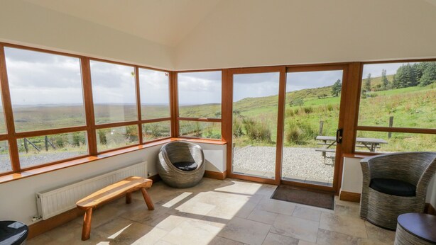 A corner room with large windows, a woven chair, a wooden bench, and a wicker chair. - Blue Stack House (Letterbarrow)