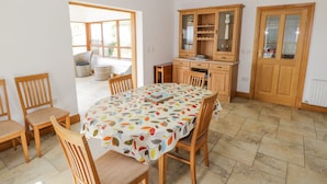 A dining area with a round table covered by a leaf-patterned tablecloth, surrounded by wooden chairs. There is a wooden cabinet with glass doors to the right, and a door with a glass panel to the right of the cabinet. - Blue Stack House (Letterbarrow)
