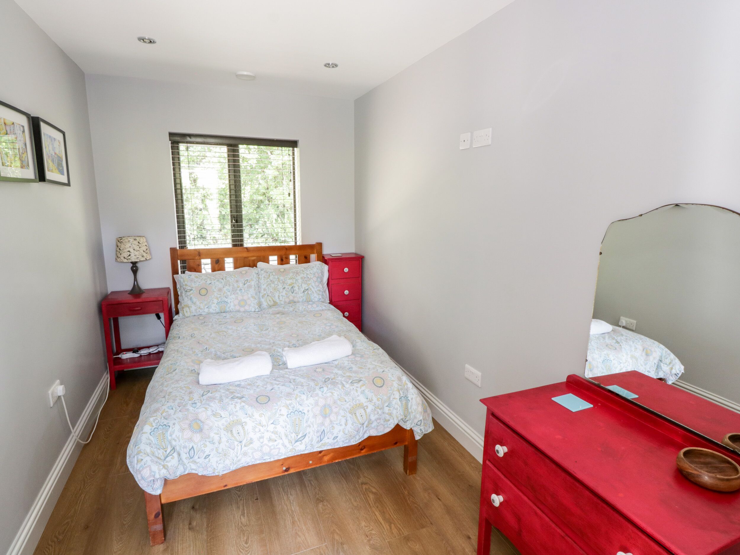 A bedroom with a wooden bed, a red nightstand, a red dresser, and a window with blinds.