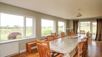 A dining room with a long table, wooden chairs, and large windows offering a view of the outdoors.