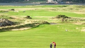 A golf course with a person in a red shirt preparing to putt, surrounded by green grass and a distant view of a town and hills.
