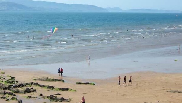 A beach with people walking and playing, and a colorful kite flying.
