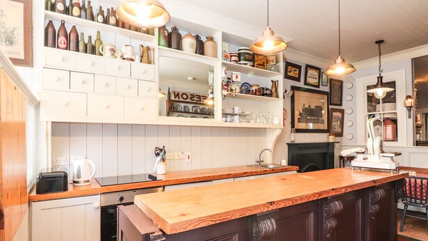 A kitchen with wooden cabinets, a central island, pendant lighting, and shelves with various kitchen items. - The Merchant's Store (Kilrush)