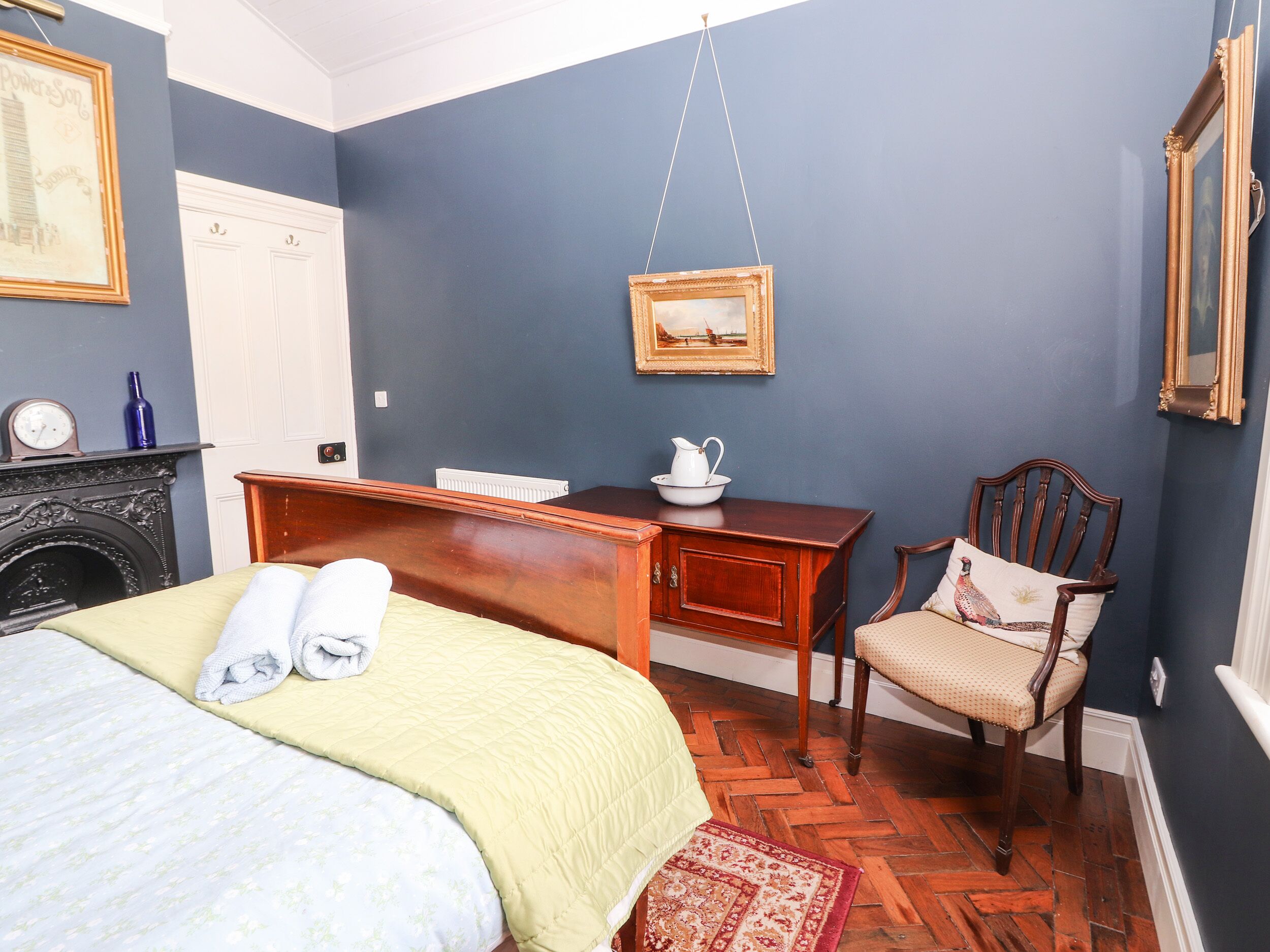 A bedroom with a bed, a desk, a chair, a fireplace, and a wall-mounted picture.
