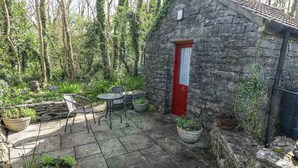 Une charmante maison en pierre avec une porte rouge, entourée d’un jardin agrémenté de plantes en pot et d’une terrasse avec des chaises en métal.