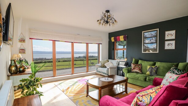 A living room with a green sofa, a red armchair, and a wooden coffee table. There is a large window with a view of a lake and mountains. - Millers Lane House (Muff)