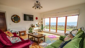 A living room with a red and green sofa set, a wooden coffee table, a TV mounted on the wall, and a large window with a view of the sea. - Millers Lane House (Muff)