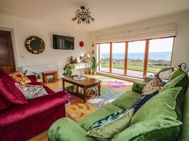 A living room with a red and green sofa set, a wooden coffee table, a TV mounted on the wall, and a large window with a view of the sea.