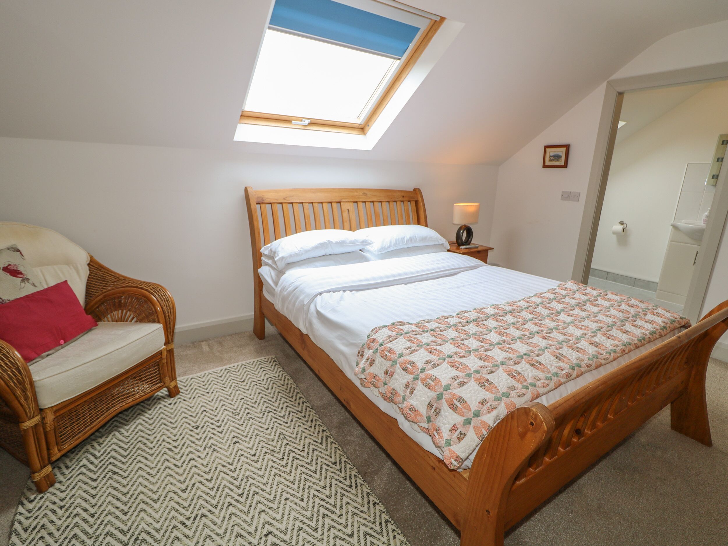 Een slaapkamer met een houten bed, een rieten stoel, een klein nachtkastje met een lamp, een dakraam en een deur.