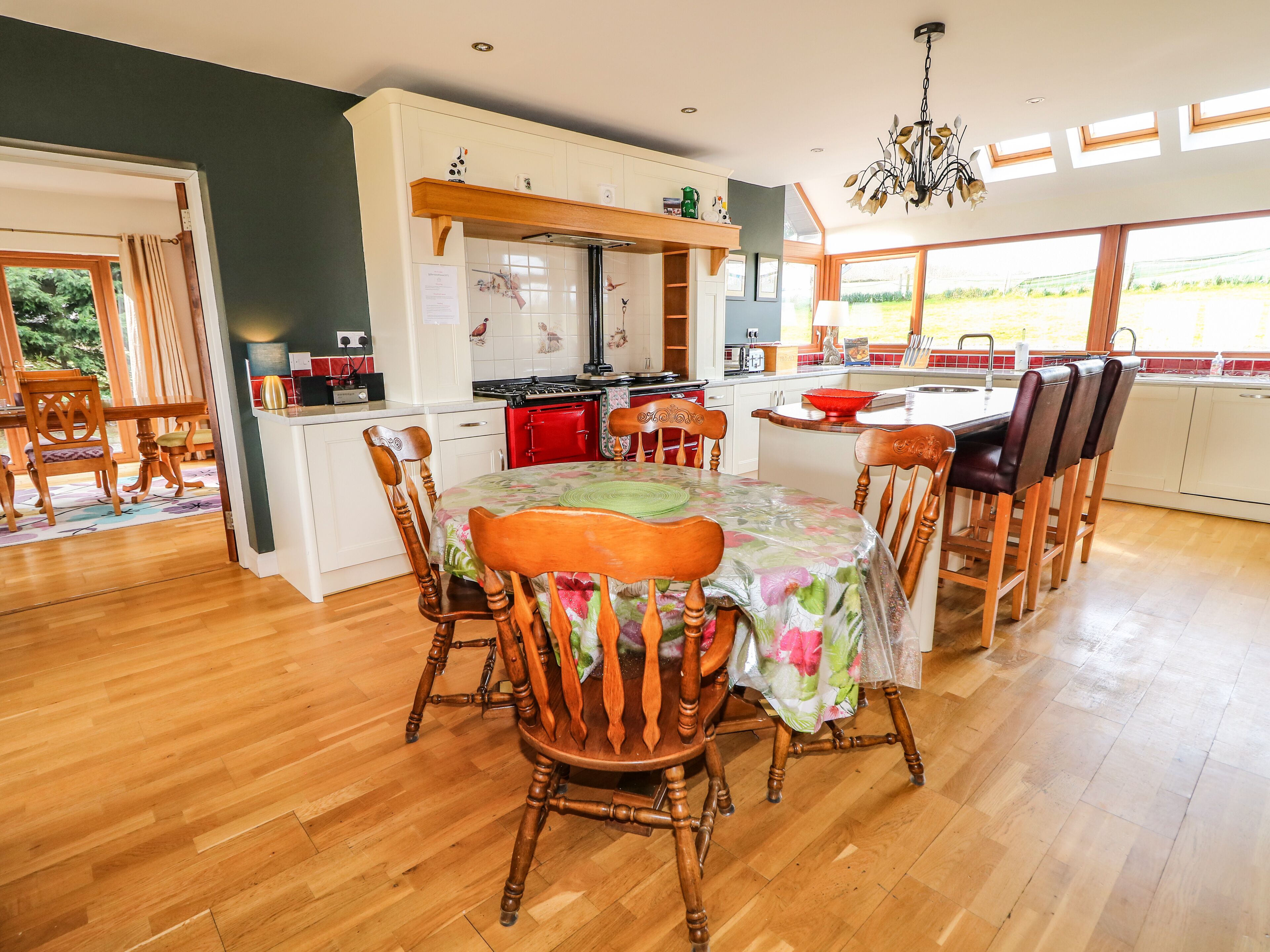 A spacious kitchen with wooden flooring, a dining area with a floral tablecloth, and large windows offering a view of the outdoors.