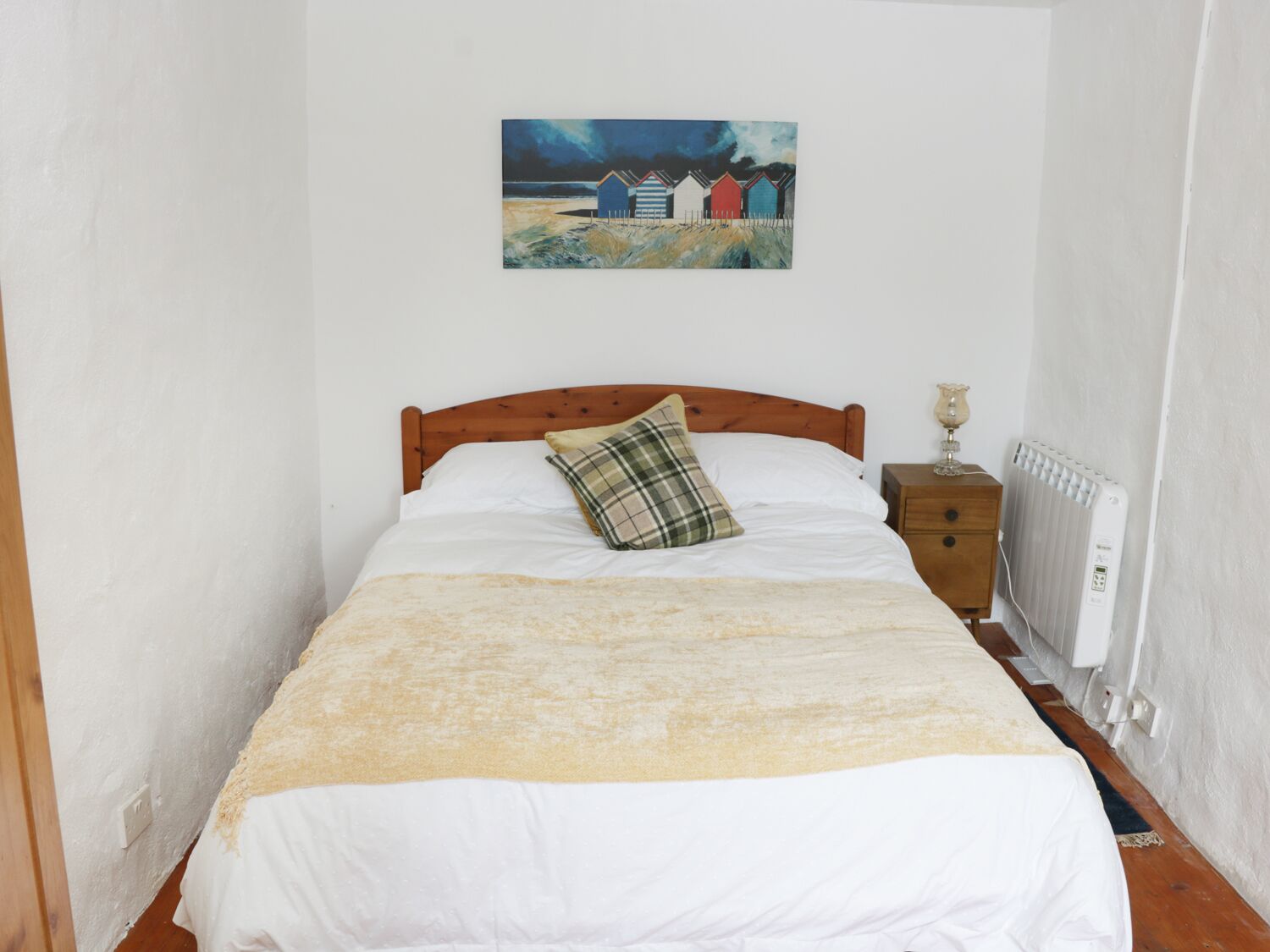 Una habitación pequeña con una cama individual, una mesita de noche, un radiador y un cuadro con cabañas en la playa.