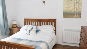 Een slaapkamer met een houten bed, wit beddengoed, een blauw-wit geruit kleed, een nachtkastje met een lamp, een wandlamp en een radiator.