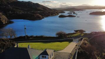 A coastal house with a paved driveway, a lawn, and a view of the sea and hills.