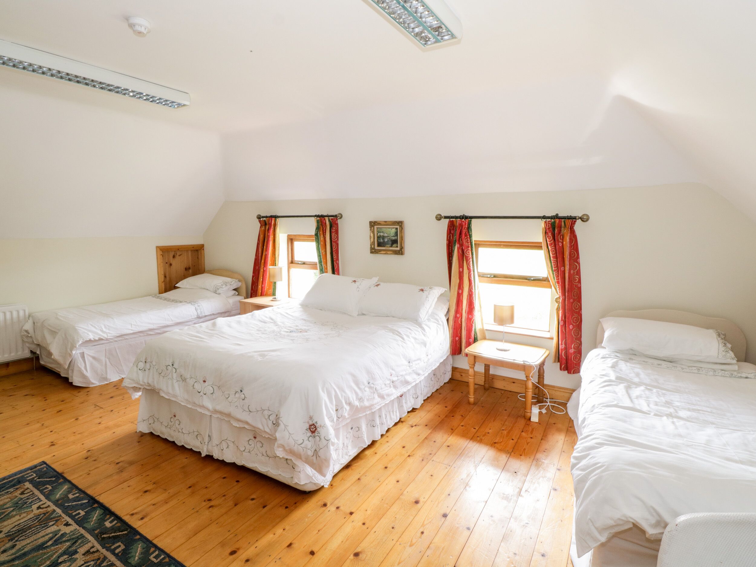 A room with two single beds, a larger bed, wooden flooring, a window with curtains, a radiator, and a ceiling light fixture.