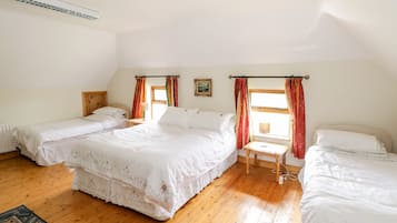 A room with two single beds, a larger bed, wooden flooring, a window with curtains, a radiator, and a ceiling light fixture.